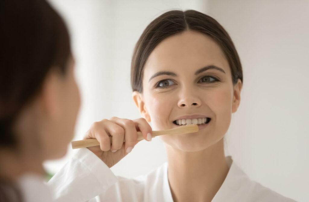 A woman brushing her teeth before bathroom mirror using soft-bristled toothbrush to care for her new dental implant.