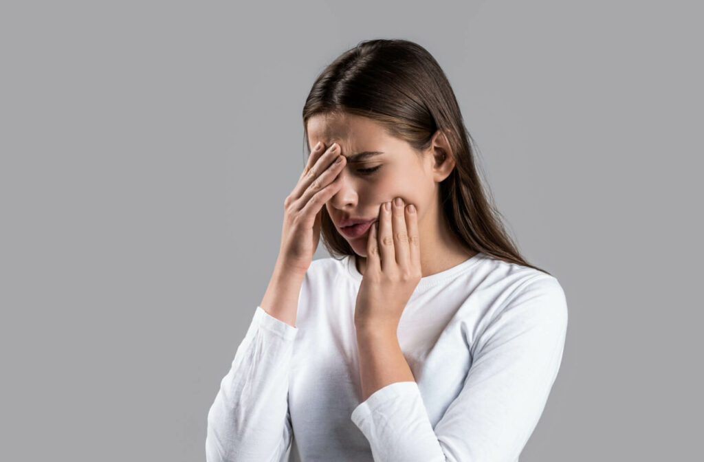 Woman in white top holding hands to forehead and cheek in pain.