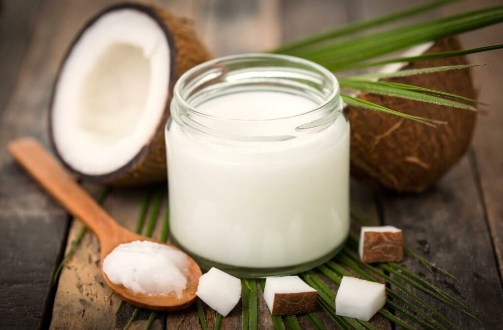 Jar of white coconut oil on a table in front of coconut pieces with a spoon of coconut oil (soft buttery solid) next to it.