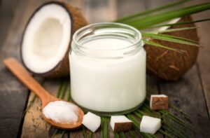 Jar of white coconut oil to whiten teeth on a table in front of coconut pieces with a spoon of coconut oil (soft buttery solid) next to it.