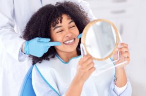 Woman with dark curly hair who was vaping before holding up mirror to bright white smile as someone with gloves stands behind and points at the smile.
