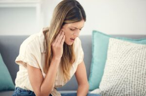 Woman with long hair sitting on a sofa and holding hand to side of her face in pain due to tooth infection