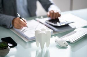 Dentist at a desk writing in a binder and tapping on a calculator with a model of a tooth in the foreground to calculate the cost of teeth cleaning