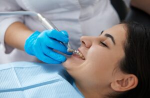 Young woman with dark hair wearing blue dental bib lying down and getting teeth cleaning by a blue gloved hand.