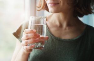 A close-up image of an adult in front of a window, holding a glass of water with fluoride in it and smiling.